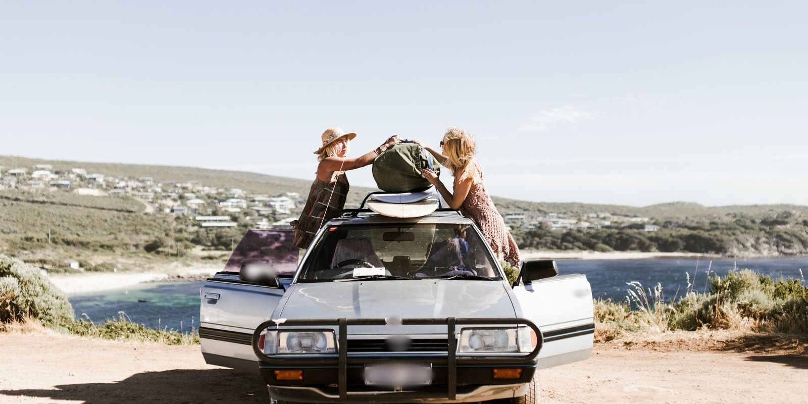 Duas mulheres colocando uma bolsa de viagem no bagageiro do teto de um carro diante de uma enseada