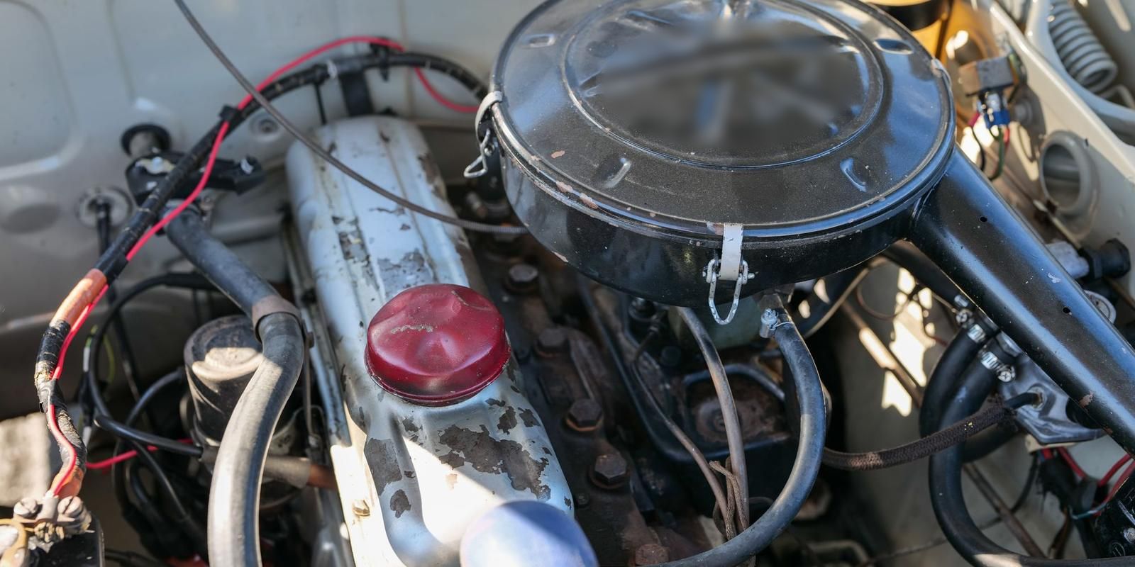 Compartimento do motor de um carro antigo visto de cima, com tampa de válvulas, mangueiras e filtro de ar