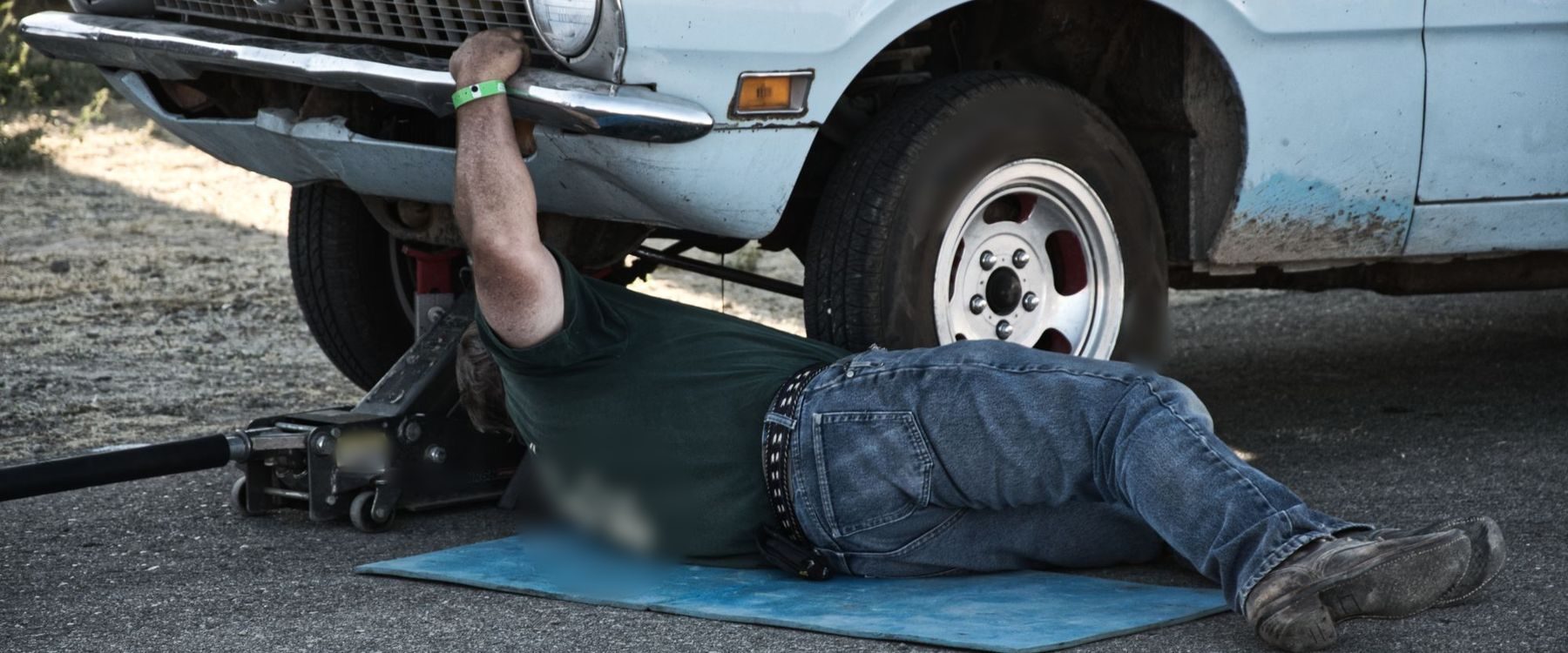 Homem deitado numa esteira azul, trabalhando sob a frente de um carro antigo erguido por macaco hidráulico